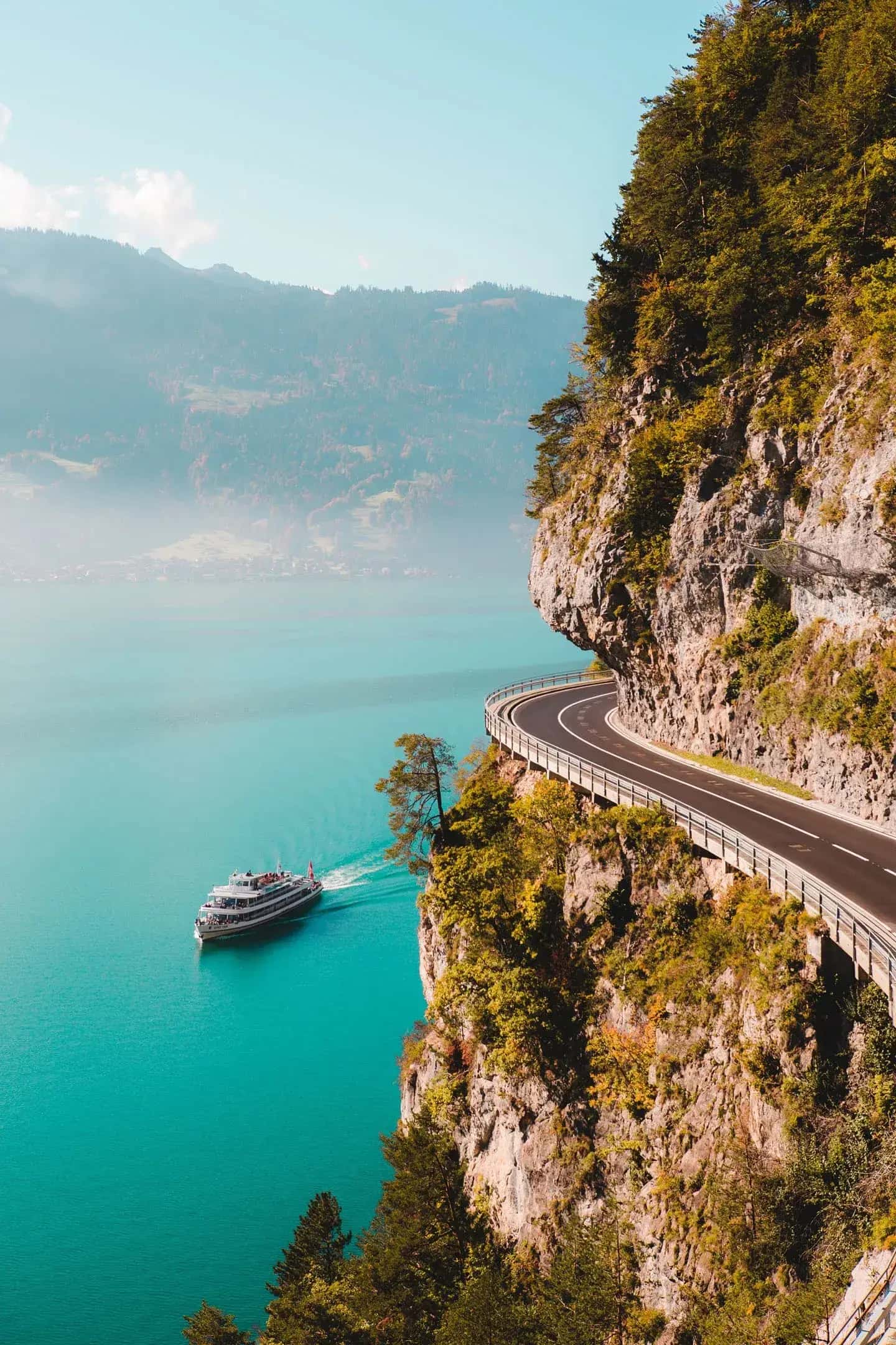 Sightseeing boat on a turquoise lake beneath a road carved into a forested cliffside