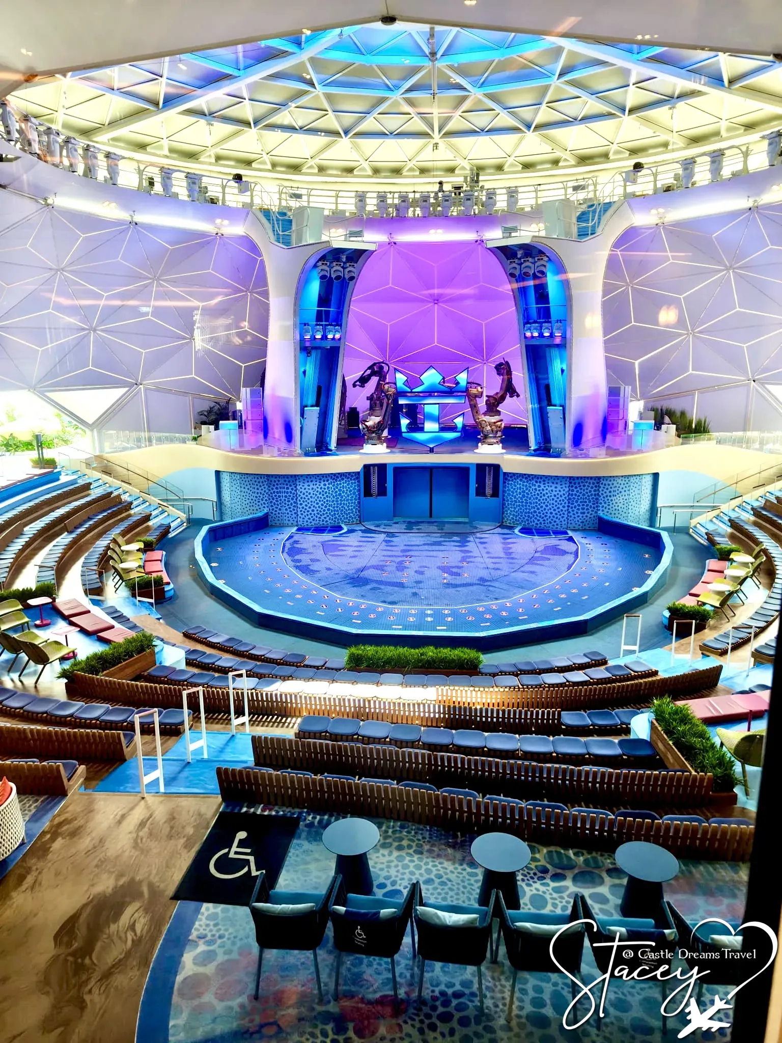 An indoor pool beneath a glass dome, lit purple, with tiered seating and a bar aboard a Royal Caribbean ship