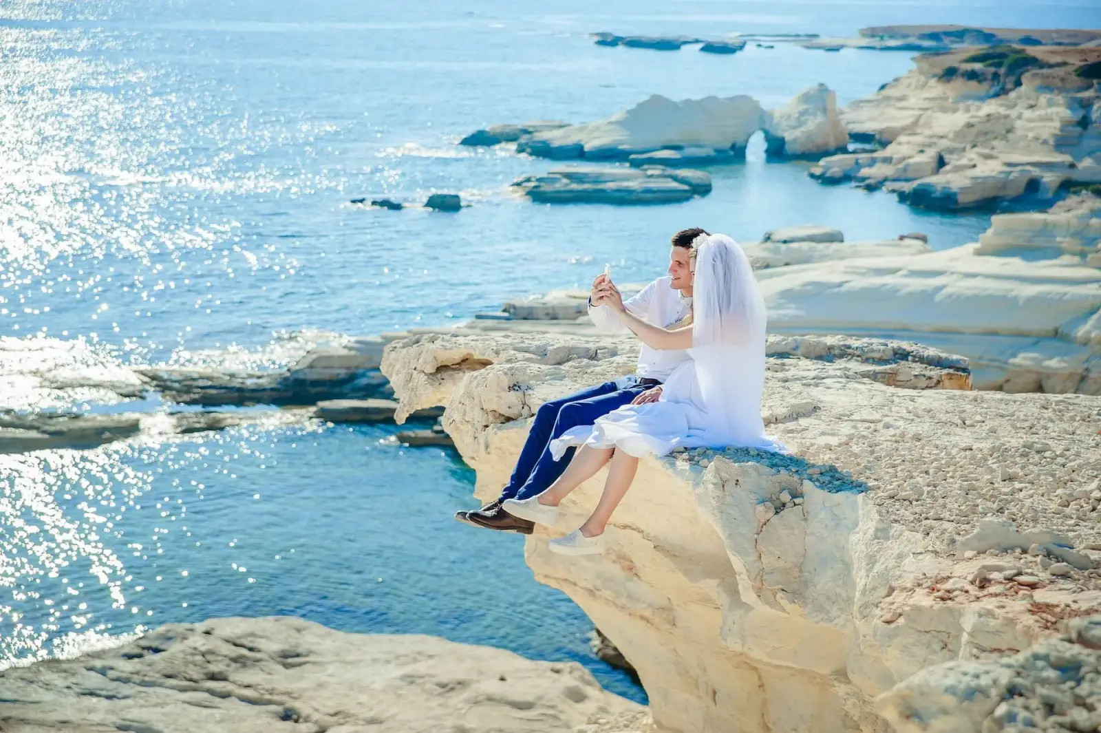 A bride and groom sitting together on white coastal rocks above a sparkling blue sea