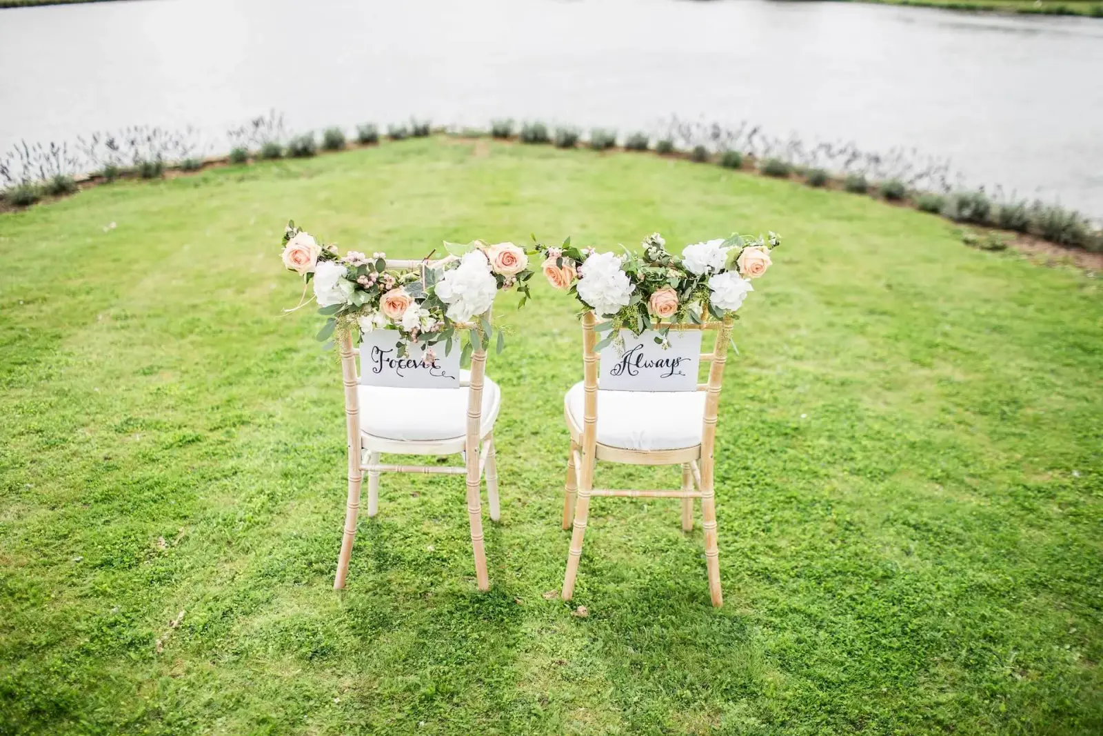 Two flower-decorated ceremony chairs labeled Forever and Always on a waterside lawn